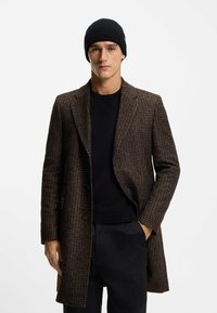 Ruskea houndstooth-takki, jossa on rakenteellinen muotoilu, kontrastivärinen musta neule alla ja sovitettu musta neulottu pipo. Sileä pinta, räätälöity istuvuus.