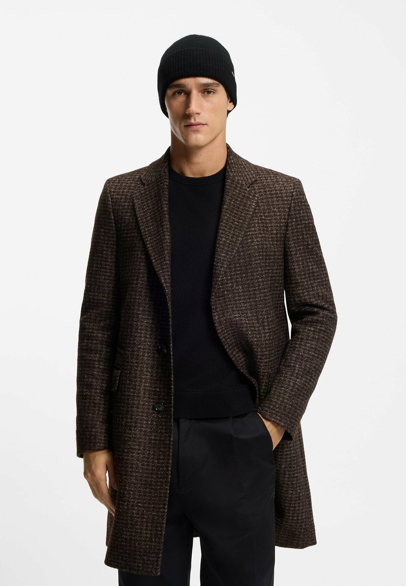 Ruskea houndstooth-takki, jossa on rakenteellinen muotoilu, kontrastivärinen musta neule alla ja sovitettu musta neulottu pipo. Sileä pinta, räätälöity istuvuus.