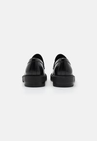 Chaussures en cuir noir avec une finition lisse, des bouts arrondis et un talon bas. Elles présentent des coutures sur les côtés et un design minimaliste.