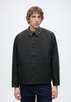 Junger Mann mit kurzen lockigen Haaren, der eine schwarze, geknöpfte Jacke mit kariertem Kragen und dunkelblaue Jeans trägt, steht vor einem einfarbigen weißen Hintergrund.