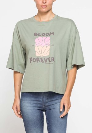 Donna che indossa una maglietta verde chiaro larga con grafica floreale rosa e beige e testo "Bloom Forever" abbinata a jeans blu.