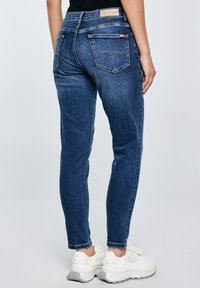 Sötétkék farmernadrágot és fehér, vaskos sportcipőt viselő személy hátulnézetből, egyszínű világos háttér előtt.