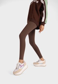 Leggings marron associés à un sweat-shirt surdimensionné marron avec des rayures vertes et blanches. Les chaussures comprennent des baskets beiges avec des accents violets.