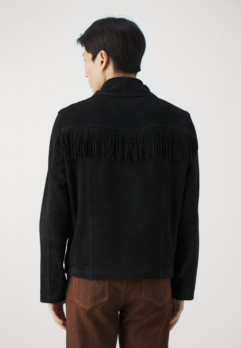 Veste en daim noir avec des détails en franges dans le dos, dotée d'un col et de manches longues. Portée avec un pantalon marron.