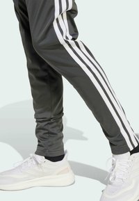 Pantaloni sportivi grigi con strisce laterali bianche, caratterizzati da una vestibilità aderente. Abbinati a sneaker bianche con dettagli testurizzati e lacci.