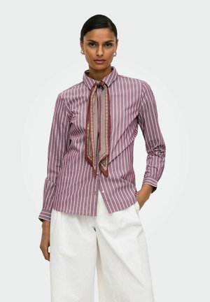 Femme portant un chemisier rayé vertical violet et blanc avec une cravate brune à motif et un pantalon blanc à jambes larges, debout, une main dans la poche.