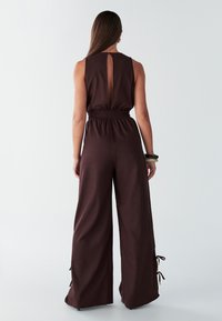 Brown sleeveless jumpsuit with a back keyhole, elastic waistband, and wide legs featuring tie accents at the hems. Smooth fabric texture.