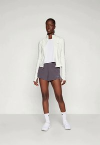 Weißer Sportjacke mit glatter Textur, kombiniert mit einem weißen T-Shirt und dunkelgrauen Shorts. Weiße Sneakers mit dezentem Branding.