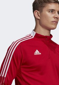 Rote sportliche Half-Zip-Jacke mit weißen Akzenten. Mit Adidas-Logo auf der Brust, drei weißen Streifen auf den Schultern und strukturiertem Gewebe.