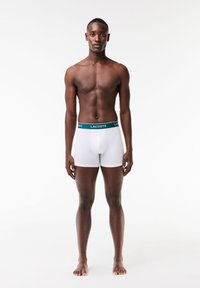 White boxer briefs with Lacoste branding on a green waistband, featuring a fitted design and smooth texture. Model stands barefoot.