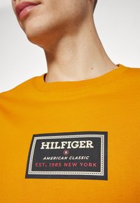 Osoba v hořčicově žluté košili s černým obdélníkovým logem s nápisem "HILFIGER American Classic EST. 1985 NEW YORK."