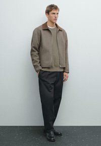 Brown wool blend bomber jacket with a brown collar, paired with a grey ribbed sweater and black wide-leg trousers, finished with black shoes.