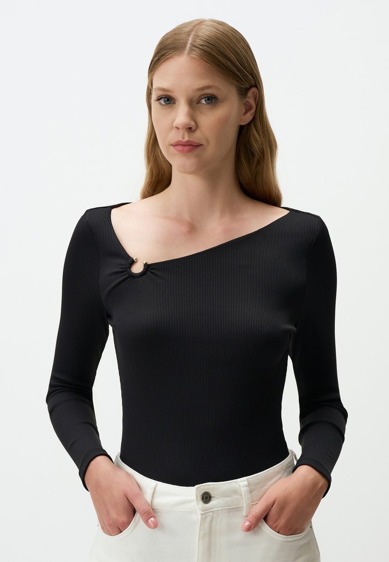 Zwarte geribbelde lange mouw bodysuit met een asymmetrische halslijn en ronde hardware details op de schouder, gecombineerd met witte hoge taille broeken.