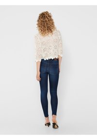 Weißes gehäkeltes Oberteil mit drei Viertel-�Ärmeln, kombiniert mit eng anliegenden dunkelblauen Jeans. Das Model trägt schwarze Absatzsandalen.
