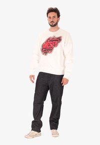 Uomo con barba che indossa una felpa bianca con testo grafico rosso, pantaloni scuri e sneakers bianche, in piedi contro uno sfondo neutro.