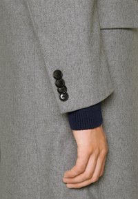 Manche de manteau en laine grise avec cinq boutons noirs, poignet côtelé bleu marine et texture lisse, mettant en avant des lignes épurées et un design structuré.