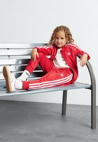 Chándal rojo de tela suave con rayas blancas, combinado con zapatillas blancas y calcetines estampados, sentado en un banco de metal.