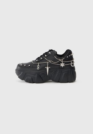 Sort chunky platform-sneaker med sølvnitter og hængende sølvcharms i kæder, herunder en dolk, sol, måne og uendelighedssymboler.