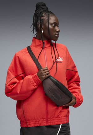 Person med flettet hår iført en rød Puma-jakke og holder en sort Puma-mavebæltetaske over brystet på en grå baggrund.