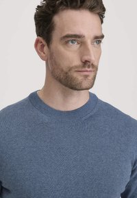 Blauer Strickpullover mit Rundhalsausschnitt, geripptem Kragen und glatter Textur. Nahaufnahme des Oberkörpers und Gesichts eines Models.