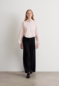 Camicia button-up corta di un rosa chiaro, con maniche lunghe, abbinata a pantaloni larghi neri. Il modello è in piedi su uno sfondo neutro.