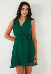 Groene mouwloze wikkeljurk met een aansluitende taille, strikceintuur en vloeiende rok. Gemaakt van lichtgewicht stof met een zachte textuur.