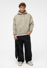 Beige Kapuzenpulli mit einer Vorderentasche, kombiniert mit locker sitzenden schwarzen Jeans. Das Model hält eine schwarze Tragetasche mit weißem Text. Die Sneakers sind hellgrau.