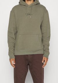 Bluza z kapturem w kolorze oliwkowej zieleni, z kapturem regulowanym sznurkiem, przednią kieszenią i wyszywanym logo. Miękka tkanina z prążkowanymi mankietami i dolnym ściągaczem. Noszona z brązowymi spodniami.