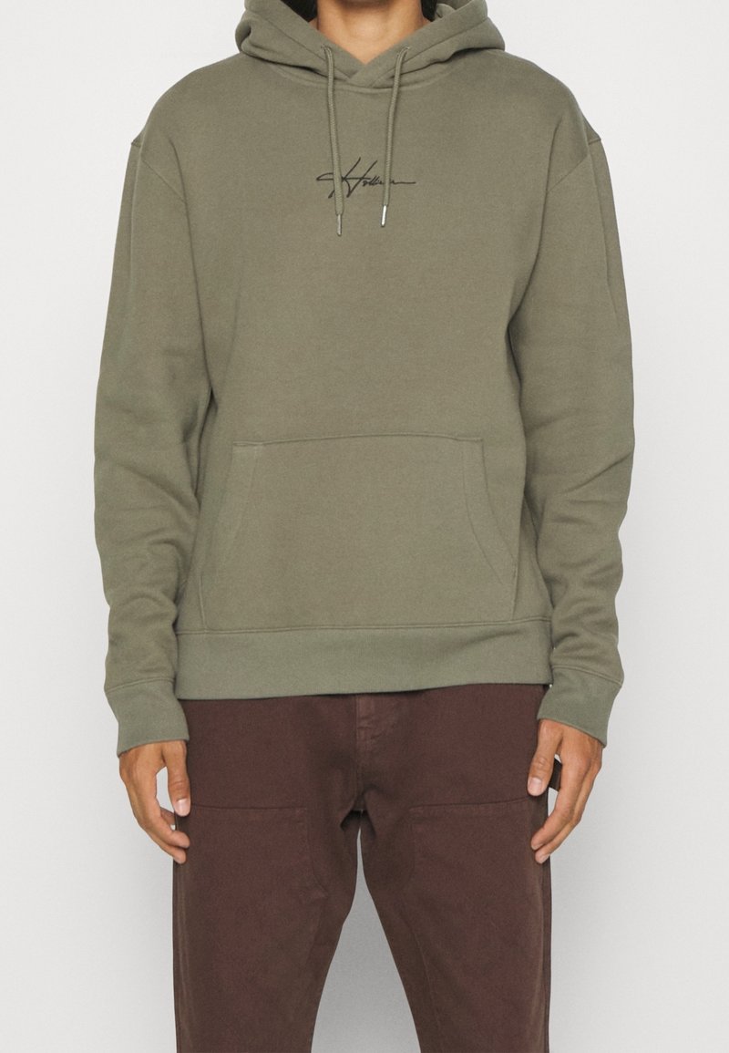 Bluza z kapturem w kolorze oliwkowej zieleni, z kapturem regulowanym sznurkiem, przednią kieszenią i wyszywanym logo. Miękka tkanina z prążkowanymi mankietami i dolnym ściągaczem. Noszona z brązowymi spodniami.
