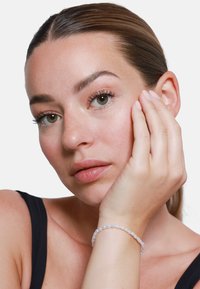 Bracelet en perles transparentes sur le poignet, composé de perles arrondies dans des tons clairs. Le sujet a les cheveux raides et un maquillage neutre.