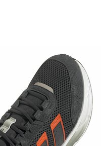 Chaussure de sport noire avec tige en mesh, accents orange et superpositions texturées. Présente des lacets noirs, une languette grise et une semelle intermédiaire blanche.