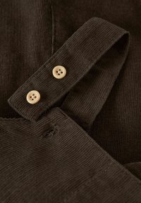 Pruunist korduroist kanga, kus on silmapaistvad ribid, millel on kaks heledat nuppu paelal, koos õmblustehnikaga ja kerge kulumisega.