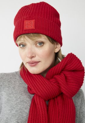 Rode geribbelde beanie met een vierkante logopatch en een bijpassende gestructureerde sjaal, stijlvol om de nek gewikkeld, over een grijze trui.