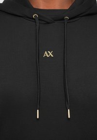 Gros plan sur l'avant d'un sweat à capuche noir avec le logo doré « AX » centré sous des cordons noirs avec embouts et œillets dorés.