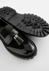 Le scarpe in pelle nera presentano una finitura lucida in vernice con dettagli brogue, una fascia in suede e pon-pon decorativi, posizionate su una suola in gomma texture.