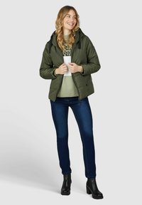 Doudoune verte avec capuche, fermeture éclair à l'avant, aspect légèrement satiné, associée à un pull léger et un jean bleu foncé, complétée par des bottines noires.