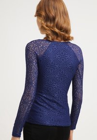 Donna con capelli rossi mossi e lunghi fino alle spalle, che indossa un top in pizzo blu navy attillato a maniche lunghe con un motivo geometrico, vista di spalle.