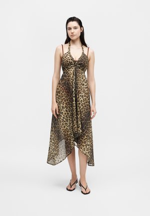 Νεαρή γυναίκα που φοράει μια αμάνικη midi φόρεμα με λεοπάρ μοτίβο, ασύμμετρο τελείωμα και μαύρα σανδάλια, στέκεται μπροστά σε ένα απλό λευκό φόντο.