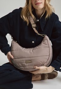 Kuriert, gepolsterte Umhängetasche in hellem Taupe mit horizontaler Steppnaht. Verfügt über einen abnehmbaren Schultergurt und ein auf der Vorderseite angebrachtes Logo-Emblem.