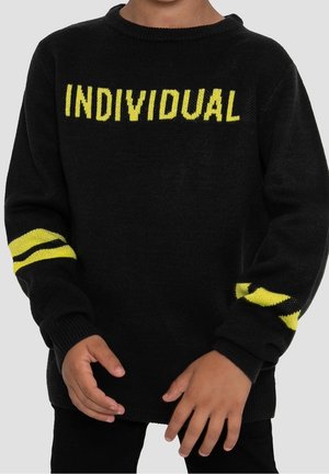 Kind draagt een zwarte trui met gele tekst "INDIVIDUAL" over de borst en gele strepen op beide mouwen, staat met ontspannen handen.