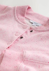 Lyserød sweatshirt med knapper foran og ribkantet krave, med en priklet tekstur og lyserøde trykknapper, samt en sort knap under kraven.