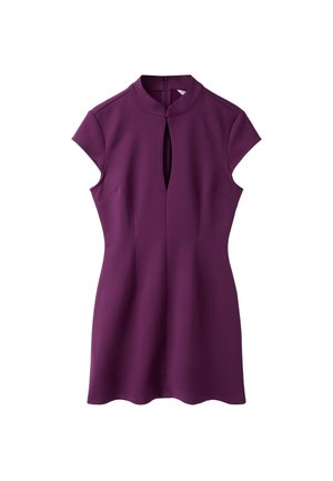 Femme debout portant une mini-robe violette à manches courtes avec une ouverture en goutte à l'avant et des chaussures slingback noires à talons hauts sur un fond gris.