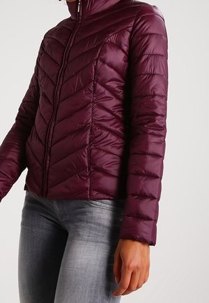 Kvinde iført en tætsiddende, quiltet vinrød jakke med lynlås foran og lysegrå jeans mod en ensfarvet baggrund.