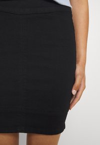 Svart denimkjol med en åtsittande design, knälång längd och synliga sömnadsdetaljer. Handen vilar på kjolens sida.