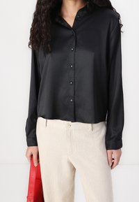 Donna che indossa una camicetta nera di raso con bottoni e pantaloni beige, mentre tiene una borsa rossa al suo fianco su uno sfondo chiaro.