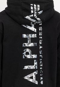 Sudadera negra con letras de camuflaje reflectantes y en negrita que dicen "ALPHA INDUSTRIES". Fabricada en un tejido suave y con capucha ajustable.