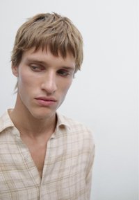 Camicia a scacchi color beige chiaro con colletto abbottonato, indossata da una persona con capelli castano chiaro disordinati su uno sfondo neutro.