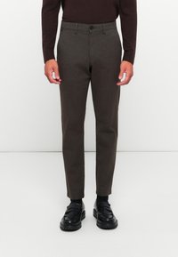 Brown wool trousers with a tapered shape, featuring a button closure and side pockets, paired with black loafers and dark socks.