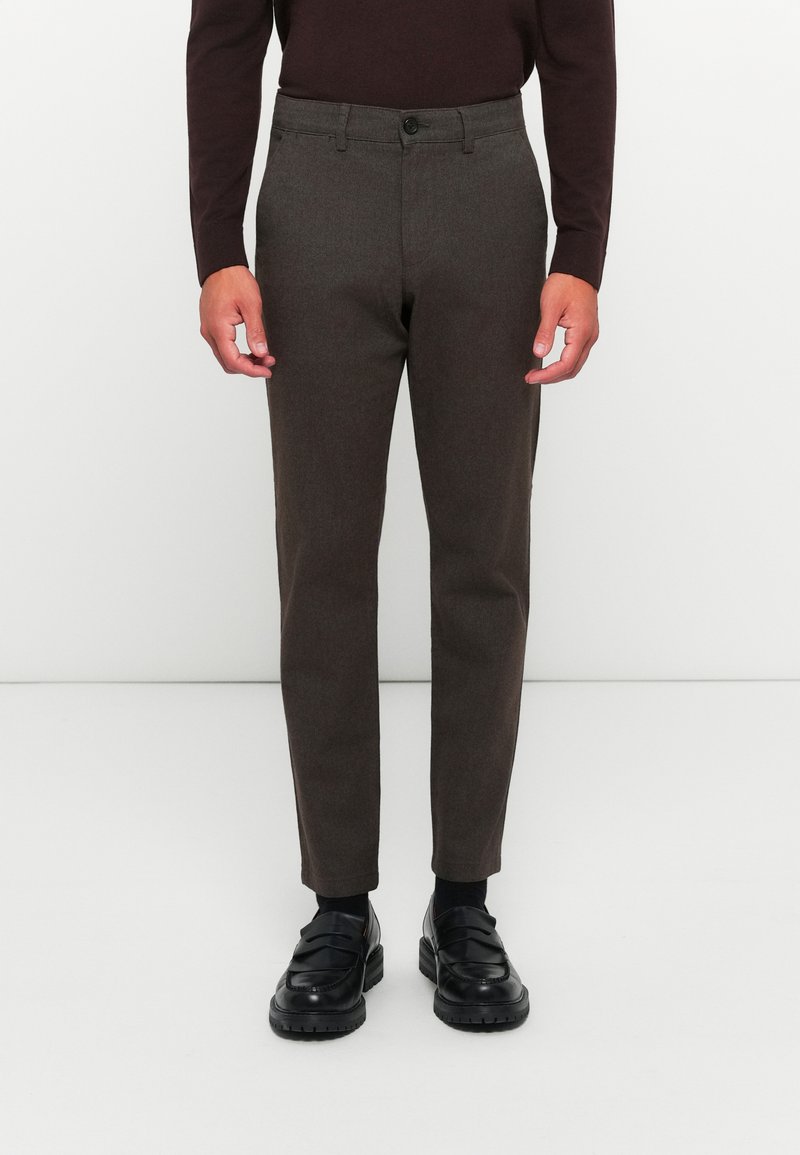 Brown wool trousers with a tapered shape, featuring a button closure and side pockets, paired with black loafers and dark socks.