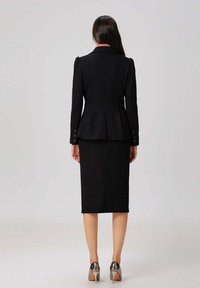 Femme de dos portant un blazer noir ajusté avec des boutons et une jupe crayon jusqu'au genou, assortie de chaussures à talons hauts argentées.